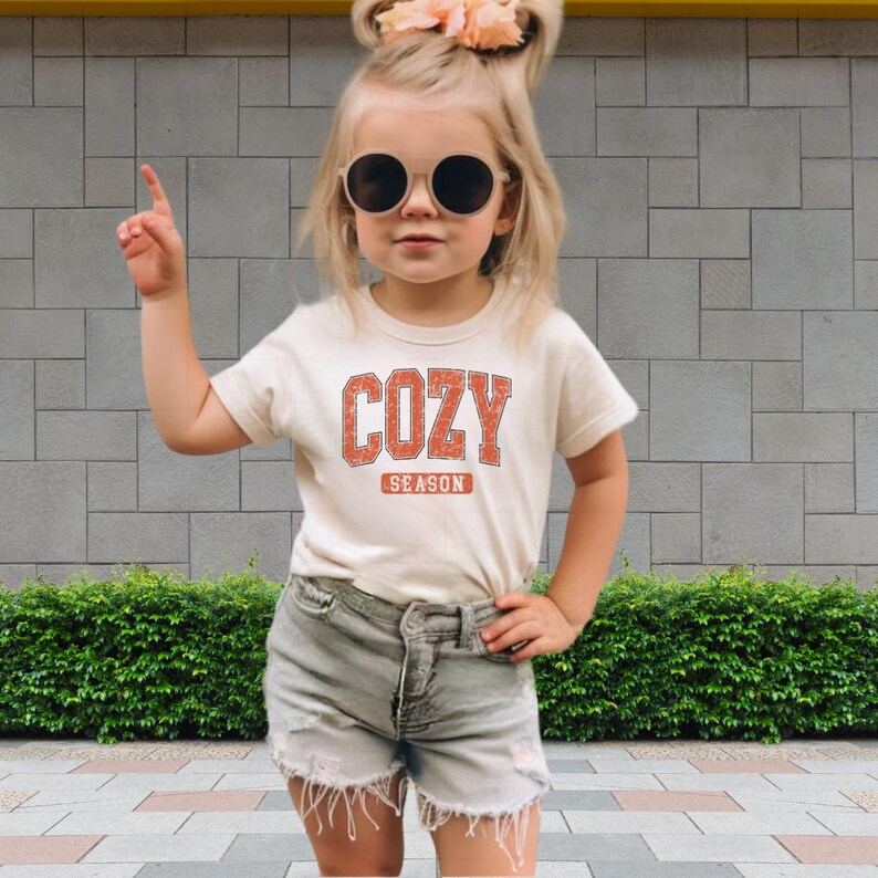 A young girl wearing a white t-shirt with the word "COZY" and sunglasses, standing in front of a brick wall and a hedge.