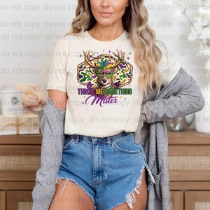 A woman wearing a white t-shirt with a colorful graphic design, denim shorts, and a gray cardigan stands in front of a wooden cabinet.