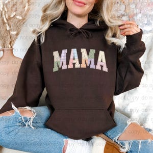 A woman wearing a brown hoodie with the word "MAMA" printed on it, sitting on the ground with her legs crossed.