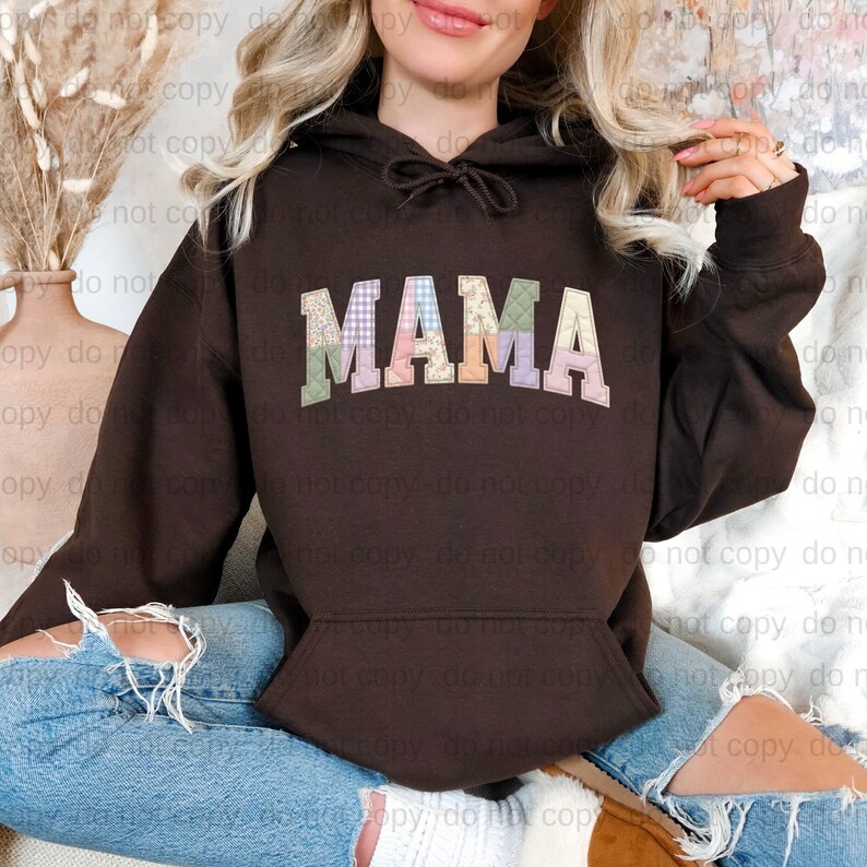 A woman wearing a brown hoodie with the word "MAMA" printed on it, sitting on the ground with her legs crossed.