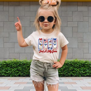 A young girl wearing a white t-shirt with a graphic design, sunglasses, and denim shorts is standing in front of a brick wall.