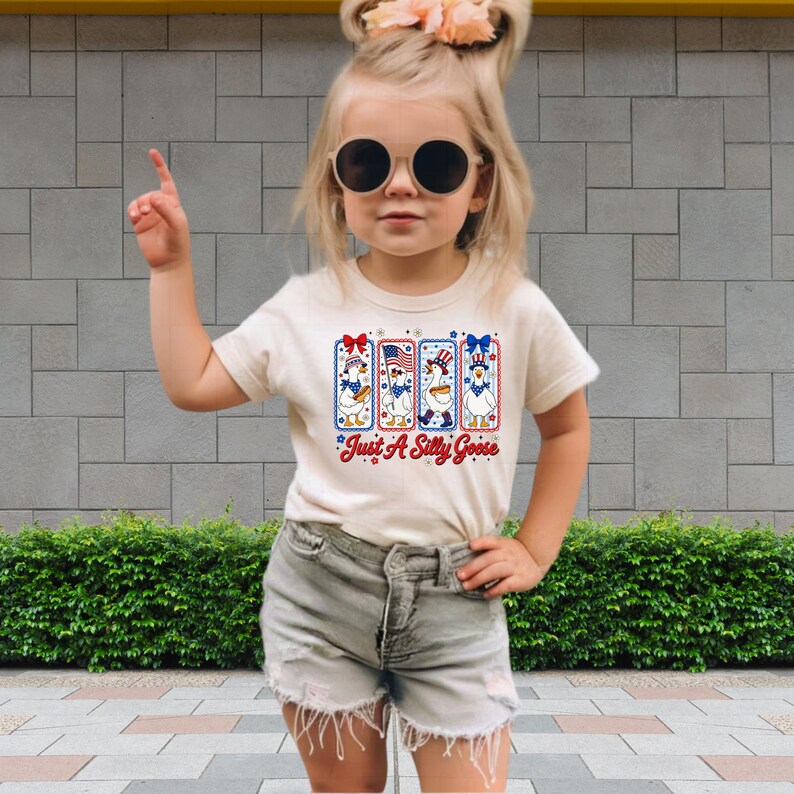 A young girl wearing a white t-shirt with a graphic design, sunglasses, and denim shorts is standing in front of a brick wall.
