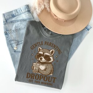 a gray t-shirt with a graphic design of a raccoon holding a drink, accompanied by the text "GENTLE PARENTING DROPOUT EST. THIS MORNING". The t-shirt is placed on top of a pair of blue jeans and a beige hat, creating a casual and laid-back outfit.