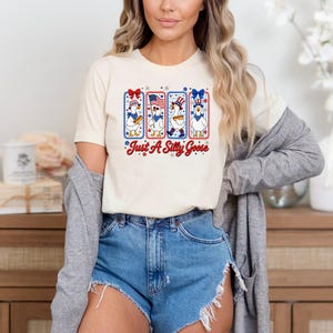 A woman wearing a white t-shirt with a graphic design and denim shorts, standing in front of a wooden cabinet.