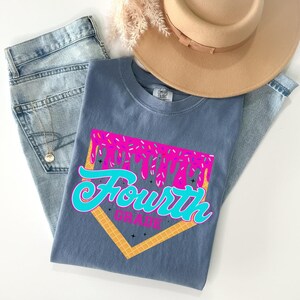 A casual outfit consisting of a gray t-shirt with a graphic design, a pair of blue jeans, and a beige hat is laid out on a white surface.