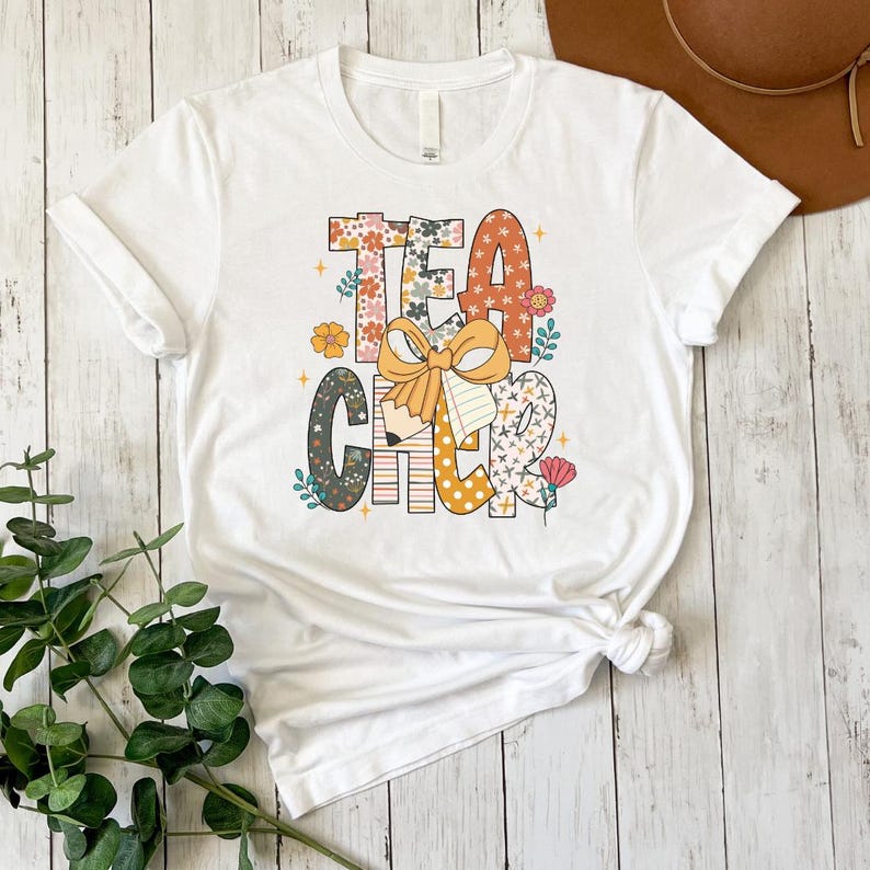 A white t-shirt with a colorful graphic design that says "Teacher" in a playful font, surrounded by various floral and decorative elements.