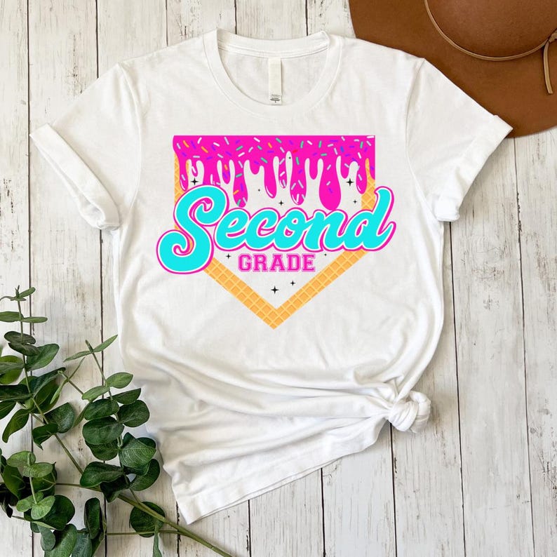 A white t-shirt with a graphic design featuring the text "Second Grade" and a dripping, colorful pattern. The t-shirt is placed on a wooden surface, with a brown hat and some greenery visible in the background.
