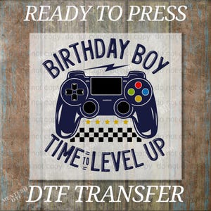 May include: A DTF transfer design featuring a navy blue video game controller with the text "BIRTHDAY BOY TIME TO LEVEL UP". The design includes a lightning bolt, stars, and a checkered pattern. The words "READY TO PRESS" and "DTF TRANSFER" are also present.