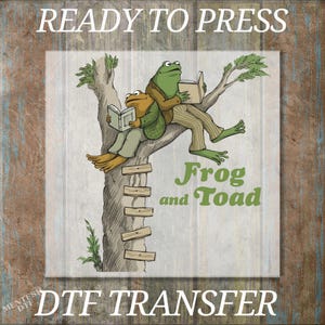 May include: A DTF transfer featuring a green frog and a brown toad sitting in a tree, each reading a book. The text reads READY TO PRESS at the top, Frog and Toad in green in the center, and DTF TRANSFER at the bottom. The design is on a wood background.