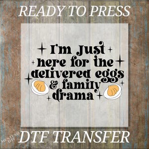 May include: A DTF transfer with the text "I'm just here for the delivered eggs & family drama." The design includes egg illustrations and the words "READY TO PRESS" and "DTF TRANSFER". The background is a wood grain pattern.