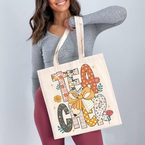 A woman is holding a large tote bag with the word "TEACHER" printed on it in a colorful, artistic style.