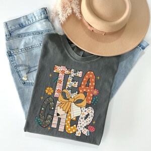 A gray t-shirt with a colorful graphic design, a beige hat, and a pair of blue jeans are arranged on a white background.