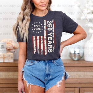 A woman wearing a black t-shirt with a graphic design and denim shorts stands in front of a wooden dresser.