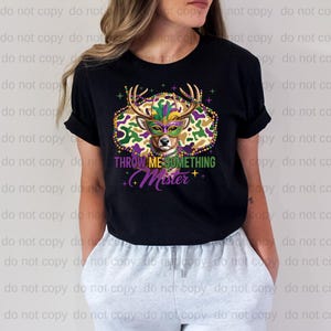 A woman wearing a black t-shirt with a colorful graphic design featuring a deer wearing a Mardi Gras mask and the text "THrowing ME SOMETHING MISTER" printed on it.