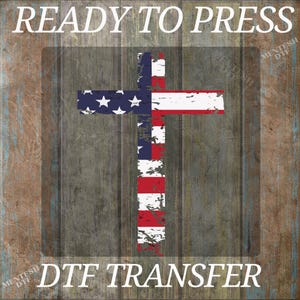 May include: DTF transfer design featuring a cross with the American flag design. The cross is red, white, and blue with stars and stripes. The words "READY TO PRESS" and "DTF TRANSFER" are in white text.