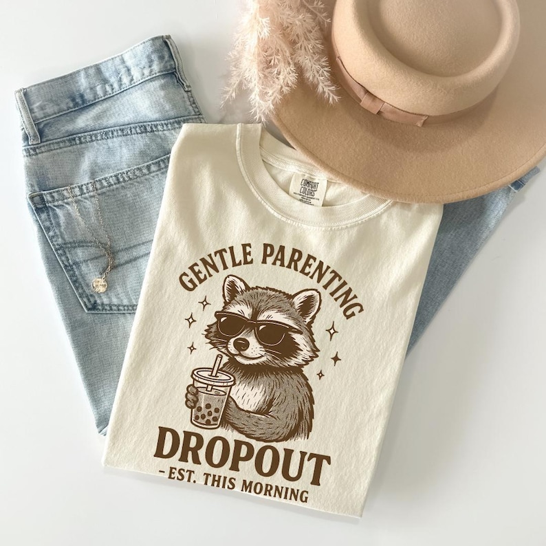 a casual outfit consisting of a white t-shirt with a raccoon graphic and text, a pair of blue jeans, and a beige hat.