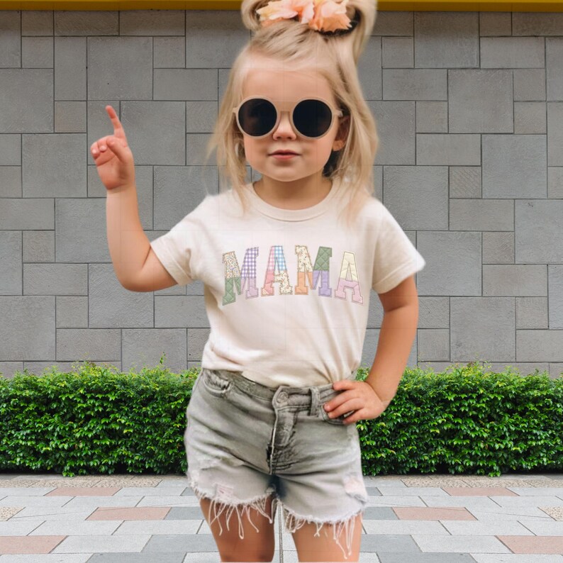 A young girl wearing a white t-shirt with the word "MAMA" and sunglasses is standing in front of a brick wall.