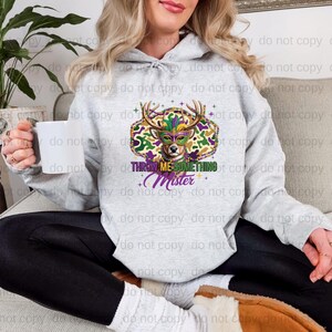 A woman wearing a grey hoodie with a colorful graphic design of a deer head and the text "throw me something" is sitting on a couch.