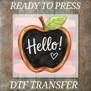 May include: A DTF transfer featuring an apple-shaped chalkboard with a brown stem and a green leaf. The center of the apple is black with the white text Hello! and a small white heart. The background consists of horizontal light pink brush strokes.