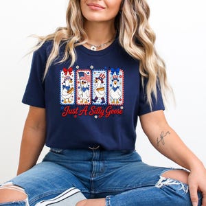 A woman wearing a navy blue t-shirt with a graphic design of four cartoon ducks and the text "Just a Silly Goose" is sitting down.
