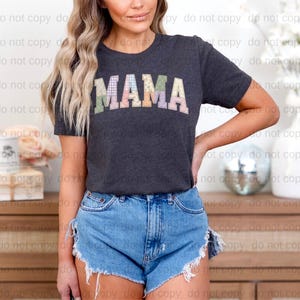 A woman wearing a black t-shirt with the word "MAMA" printed on it, paired with denim shorts, stands in front of a wooden dresser.