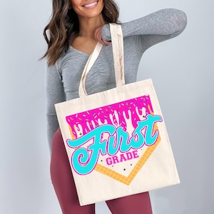 A woman is holding a large tote bag with a colorful graphic design that says "First Grade" on it.