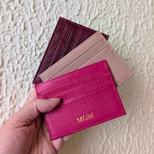 Op de afbeelding: Drie leren kaarthouders in bordeaux, beige en fuchsia. De fuchsia kaarthouder heeft het woord "MOM" in gouden letters. Elke kaarthouder heeft meerdere kaartsleuven.