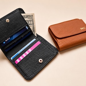 May include: A black pebbled leather wallet is open, revealing multiple card slots containing credit cards and a one-dollar bill, secured by a gold snap button. Beside it sits a closed tan pebbled leather wallet featuring the gold embossed letters MC.
