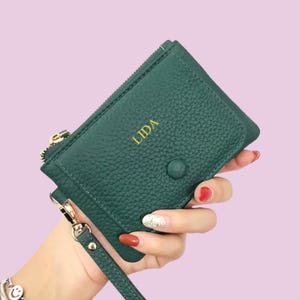 Może przedstawiać: Ciemnozielony skórzany portfel z napisem "LIDA" w złotych literach. Portfel posiada zapięcie na zamek błyskawiczny, zapięcie na zatrzask i pasek na nadgarstek. Tło jest w kolorze jasnoróżowym.