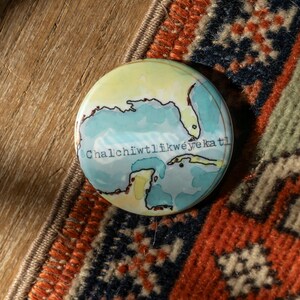 May include: A round button with a blue and green watercolor map of a coastline. The text "Chalchihuitlíkwéyé ka tl" is written on the button.