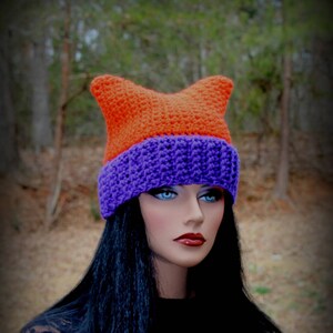 May include: A handmade crochet hat in orange and purple. The hat features a cat ear design and is worn by a mannequin with long black hair. The hat is a unique accessory for those who love handmade items.
