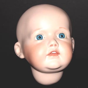 May include: A vintage doll head with a pale complexion, rosy cheeks, and a small, open mouth. The doll has blue eyes and a bald head. The head is made of porcelain and is a collectible item.