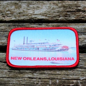 May include: A rectangular patch with a red border featuring a steamboat on a blue background. The text "NEW ORLEANS, LOUISIANA" is printed in red at the bottom of the patch. The patch is set against a weathered wooden surface.