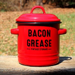 May include: A red enamel bacon grease container with a lid and handle. The container has black text that reads "BACON GREASE" and "VINTAGE STORAGE". The container is sitting on a wooden surface.
