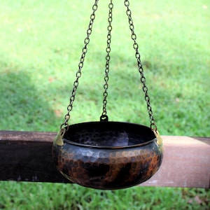 May include: A round, hammered metal bowl with a dark, mottled copper and black finish. The bowl features three brass-colored attachment points connected to three thin metal chains that meet at a single top hook for hanging display purposes.