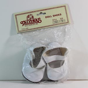 May include: A pair of white doll shoes with a strap, packaged in a clear plastic bag. The shoes are displayed under a label that reads "DOLL SHOES" and "TALLINA'S DOLL SUPPLIES INC."