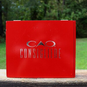 May include: A bright red box with the words "CAO CONSIGLIERE" in silver lettering. The box is sitting on a wooden surface with a green grassy background and trees.