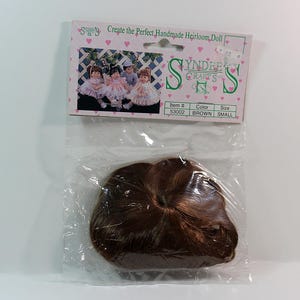 May include: A brown doll wig in a clear plastic package. The packaging includes the text "Create the Perfect Handmade Heirloom Doll" and "Syndee Crafts". The item is labeled as brown and small.