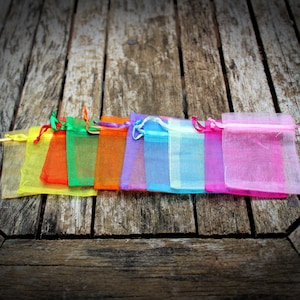 May include: A row of colorful organza gift bags in yellow, orange, green, purple, blue, and pink. Each bag has a matching drawstring closure. The bags are arranged on a weathered wooden surface.