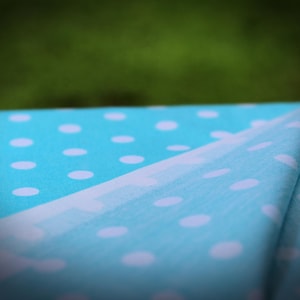 May include: A bright turquoise fabric featuring a repeating pattern of small, solid white polka dots. The material is folded over, revealing the consistent dot design across the surface of the textile, which is captured in a soft focus.