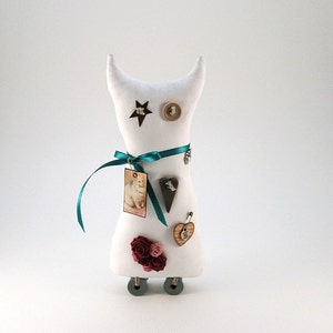 May include: White fabric doll with a cat-like face, decorated with buttons, a small picture, a heart, and a ribbon. The doll has a teal ribbon tied around its neck and is on wheels.