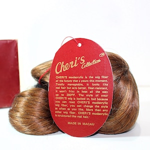 May include: A brown wig from Cheri's Collection with a red tag. The tag includes product information and the text "CHERI'S modacrylic is the wig fiber of the future". The wig is heat-resistant and can be styled.