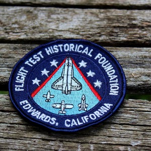 May include: A circular patch with the text "FLIGHT TEST HISTORICAL FOUNDATION EDWARDS, CALIFORNIA" in white on a navy blue background. The patch features a central design of a space shuttle and aircraft, with red and white accents.