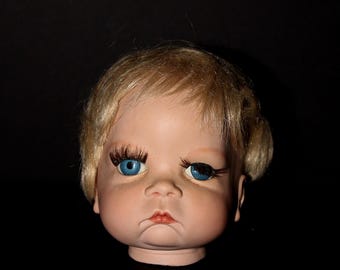 May include: A vintage doll head with blonde hair and blue eyes. The doll has long eyelashes and a slightly frowning expression. The head is set against a black background, highlighting its features.