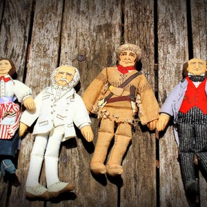 May include: Four handmade cloth dolls depicting historical figures. The dolls are dressed in period clothing, including a woman in a red and white striped apron, a man in a white suit, a frontiersman, and a man in a suit with a red vest.