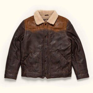 May include: A dark brown leather jacket featuring a contrasting tan leather yoke across the shoulders and chest. The jacket includes a cream-colored faux shearling collar, a front snap-button closure, and two large front pockets with snap-button flaps.