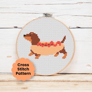 May include: A circular wooden embroidery hoop containing a cross stitch pattern of a brown dachshund dog inside a tan hot dog bun with red ketchup lines. The white fabric background features a grid of stitches. A peach scalloped label reads Cross Stitch Pattern.