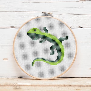 Könnte beinhalten: Kreuzstich-Stickerei mit einem grünen und dunkelgrünen Eidechsen-Design. Die Eidechse befindet sich in einem runden Stickrahmen aus Holz. Der Hintergrund ist weiß mit einem Rastermuster, und der Rahmen befindet sich auf einer weißen Holzoberfläche.