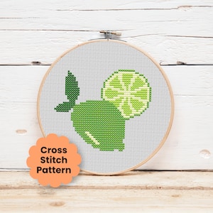 Könnte beinhalten: Ein runder Stickrahmen aus Holz mit einem Kreuzstichmuster einer Limette. Das Design zeigt eine dunkelgrüne Limette mit Blatt und ein hellgrünes Limettenscheiben-Segment auf weißem Stoff. Ein orangefarbenes, gewelltes Etikett trägt die Aufschrift Cross Stitch Pattern.