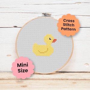 May include: A round wooden embroidery hoop containing white Aida fabric with a yellow rubber duck cross stitch pattern. The design features a small orange beak and a black eye. Text on the image reads Cross Stitch Pattern and Mini Size.
